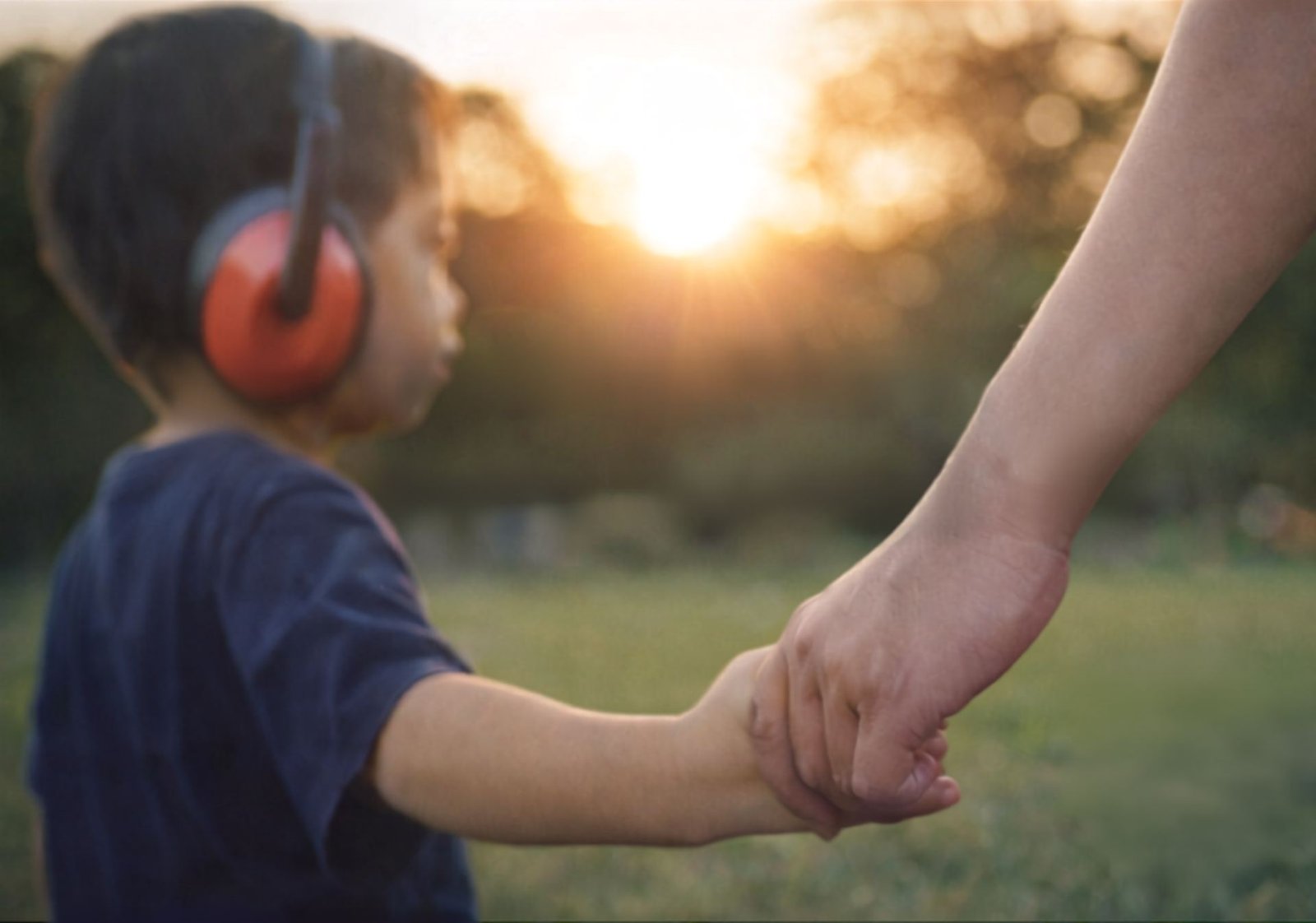 A child wearing ear defenders walks hand in hand with an adult at sunset.